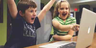Ein Junge und ein Mädchen vor einem Laptop. Das Mädchen zeigt auf den Bildschirm und der Junge hebt jubelnd die Arme. Symbolbild Gentherapie